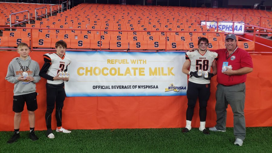 Dairy Farmer Neil Peck (far right) of Welcome Stock Farm provides chocolate milk to Schuylerville team members: nephew Hudson Peck and Hanson Peck (#3) of Welcome Stock Farm, and cousin Ryan Peck (#58) of Clear Echo Farm after the NYSPHSAA Class C State Championship game. Photo provided.